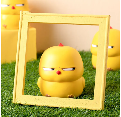 A cute, yellow chick stress ball toy with a playful expression, framed inside a yellow square frame against a grassy background.